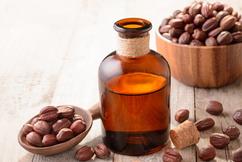 Jojoba oil one of the best hair oil options for 4c hair in bottle on wooden table next to a spoon with jojoba seeds. Behind the bottle is a wooden bowl of jojoba seeds